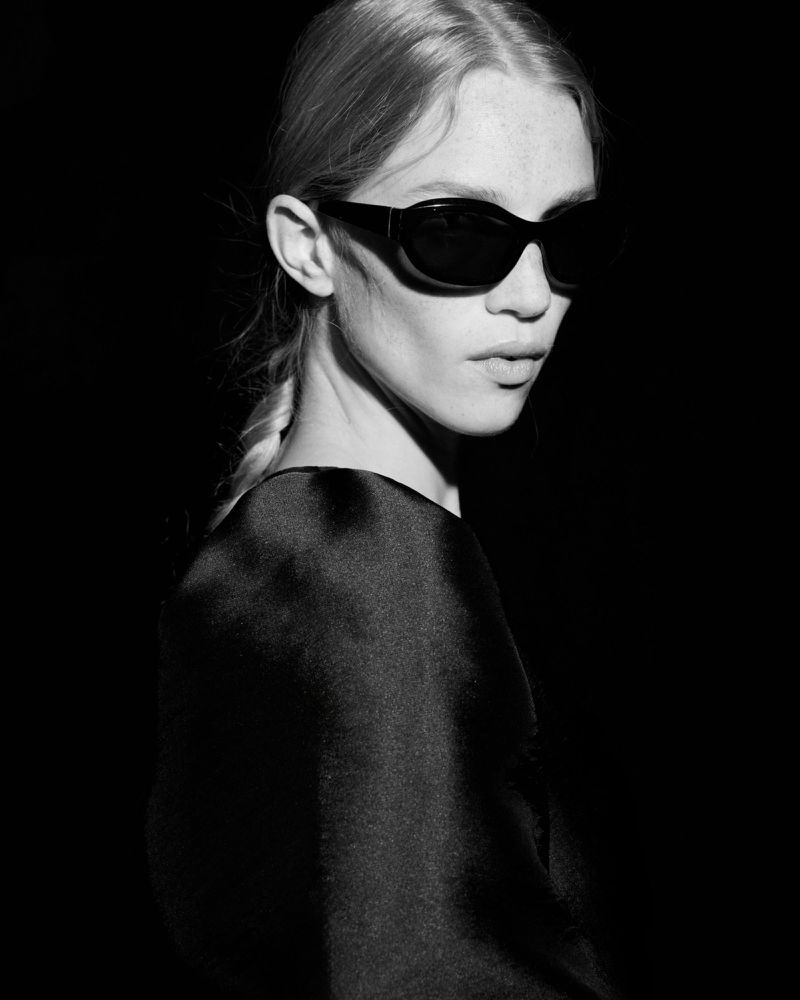 Monochrome portrait of a woman with blonde hair in a braid wearing sunglasses and a dark dress against a stark black background.