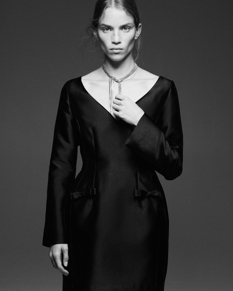 Model Rebecca Leigh Longendyke wearing a black long-sleeved V-neck dress and silver chain necklace in a studio portrait.