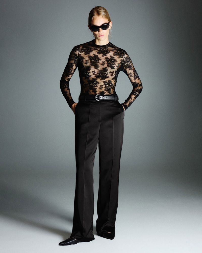 Fashion model in black lace top and black pants posing in studio with hands in pockets and wearing sunglasses.