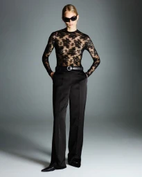 Model Rebecca Leigh Longendyke in a sheer black lace top, wide-leg trousers, and sunglasses against a grey studio backdrop.