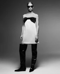 Rebecca Leigh Longendyke wearing a white strapless mini dress with a black bow and thigh-high black boots against a studio backdrop.