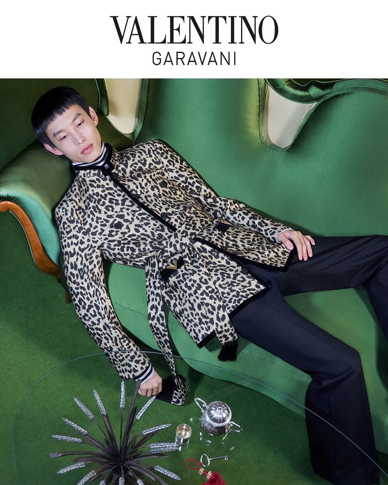 Elegant Asian man in a leopard-print jacket lounges on a green sofa with a vintage tea set, captured in a high-angle fashion shot.