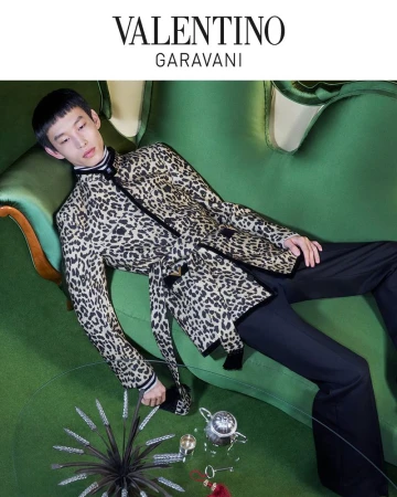 Elegant Asian man in a leopard-print jacket lounges on a green sofa with a vintage tea set, captured in a high-angle fashion shot.