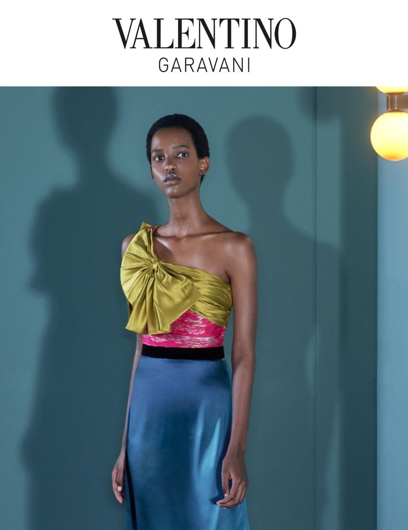 Fashion editorial shot of a Black woman in a one-shoulder satin top and blue skirt against a teal backdrop with soft lighting and shadows.