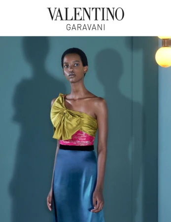 Fashion editorial shot of a Black woman in a one-shoulder satin top and blue skirt against a teal backdrop with soft lighting and shadows.