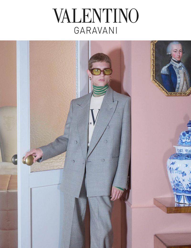Valentino fashion ad: Man poses in a gray suit next to a pink wall, white door, and blue-and-white decorative vase.