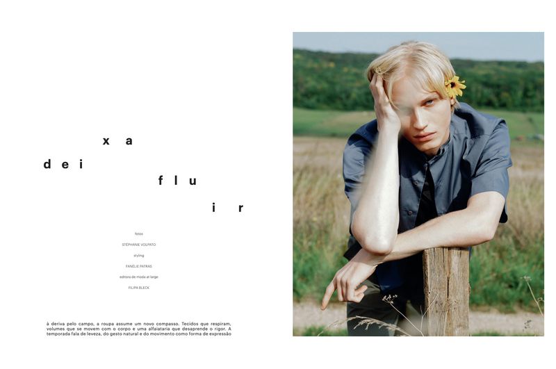 Blond male model with sunflower posing in a rural field, wearing a blue button-down shirt, leaning on a wooden post, editorial fashion.