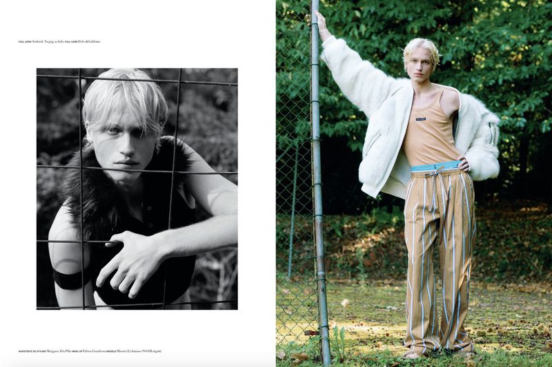Young male model poses in a tan tank top, white jacket, and striped trousers against a green leafy backdrop, half in black and white.