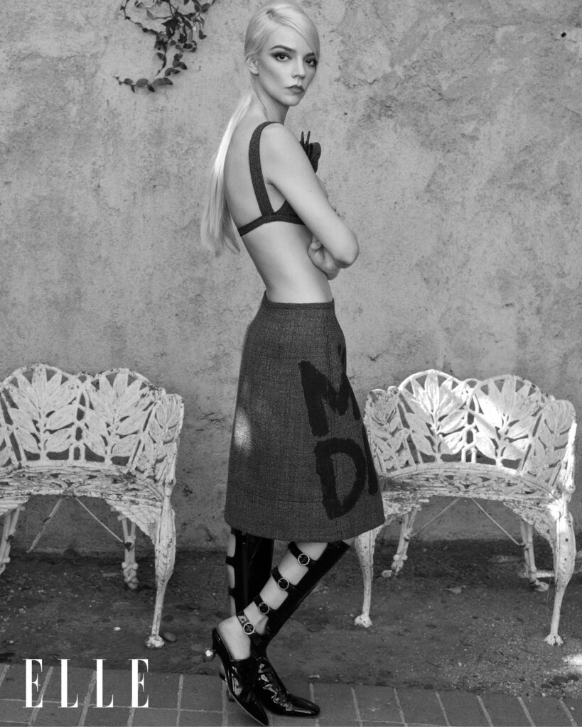 Black and white fashion portrait of a fair-skinned woman in a skirt and bra, standing in an outdoor setting with ornate iron benches.