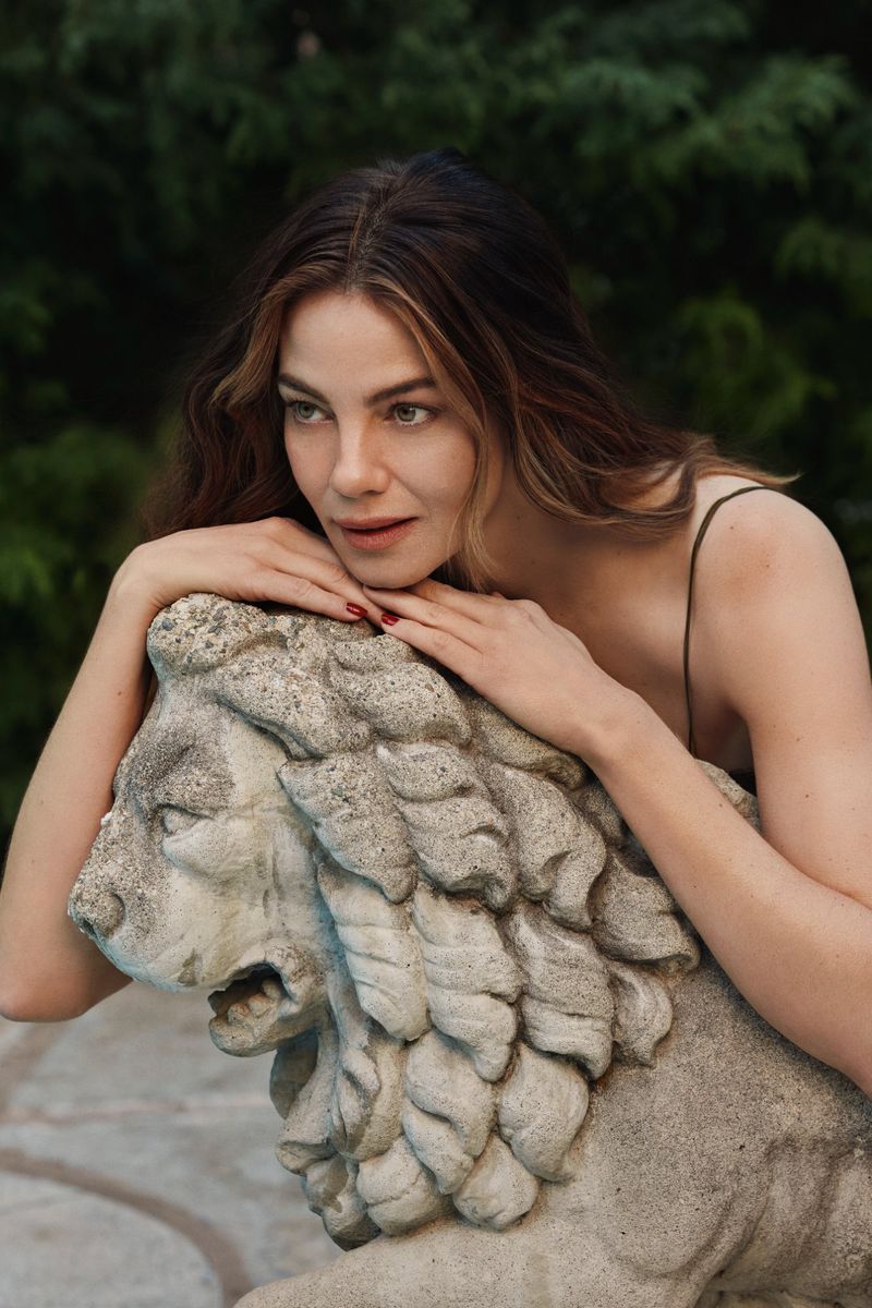 Adult woman rests her face on a stone lion sculpture outdoors, gazing to the side with lush green vegetation in the background.