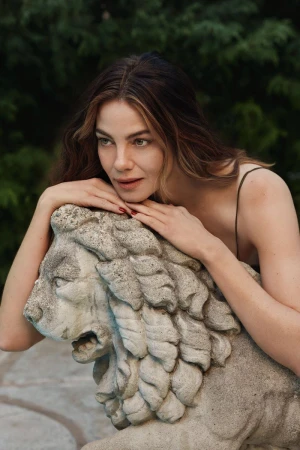 Adult woman rests her face on a stone lion sculpture outdoors, gazing to the side with lush green vegetation in the background.