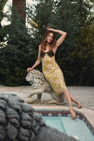 Woman in a yellow dress poses next to a lion statue by a pool surrounded by trees in an outdoor environment; fashion-style photography.