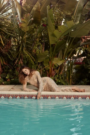 Woman in shimmering dress poses beside a pool surrounded by lush tropical foliage, creating an elegant fashion editorial shot.