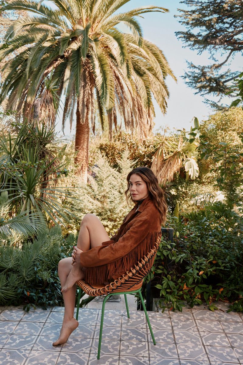 Adult female poses in a fringed jacket on a wicker chair outdoors surrounded by lush foliage and a large palm tree on a sunny day.