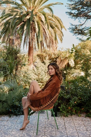 Adult female poses in a fringed jacket on a wicker chair outdoors surrounded by lush foliage and a large palm tree on a sunny day.