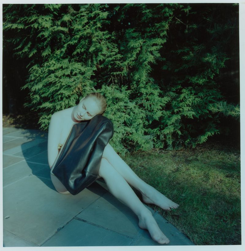 Woman with fair skin poses outdoors with a black leather bag, seated on a stone surface with a lush green tree background in natural light.
