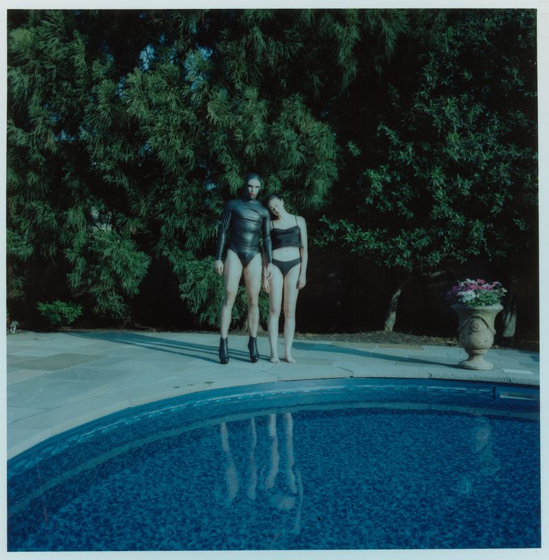Two people in swimsuits stand by a pool against a backdrop of lush greenery, captured in a low-angle outdoor setting on a sunny day.
