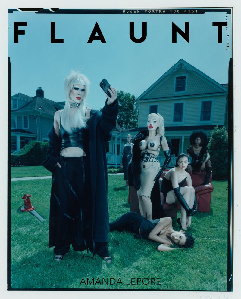 Editorial fashion shot of Amanda Lepore with several other models outdoors in front of a house and green lawn with an axe prop.
