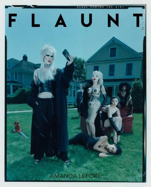 Editorial fashion shot of Amanda Lepore with several other models outdoors in front of a house and green lawn with an axe prop.