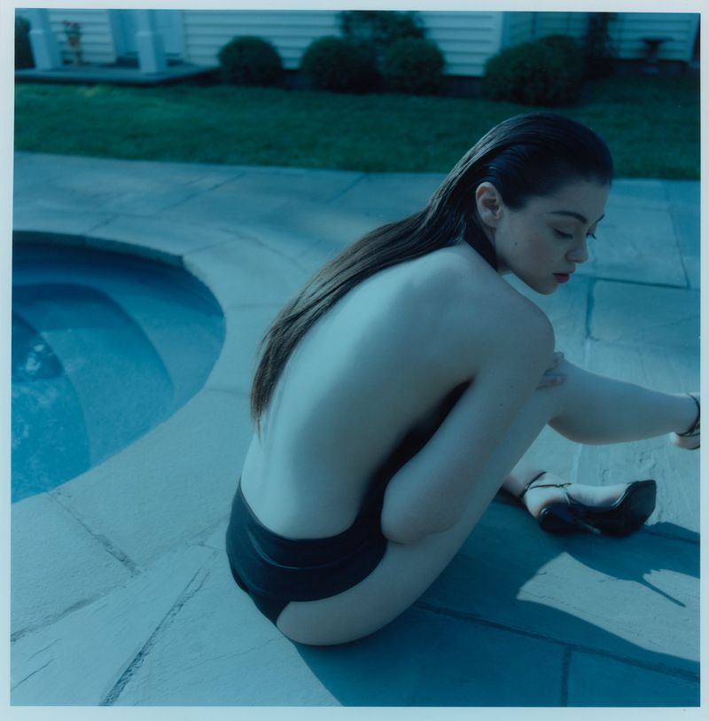 Woman crouching poolside on stone patio tiles wearing black bikini bottoms and high heels under cool blue-toned light.