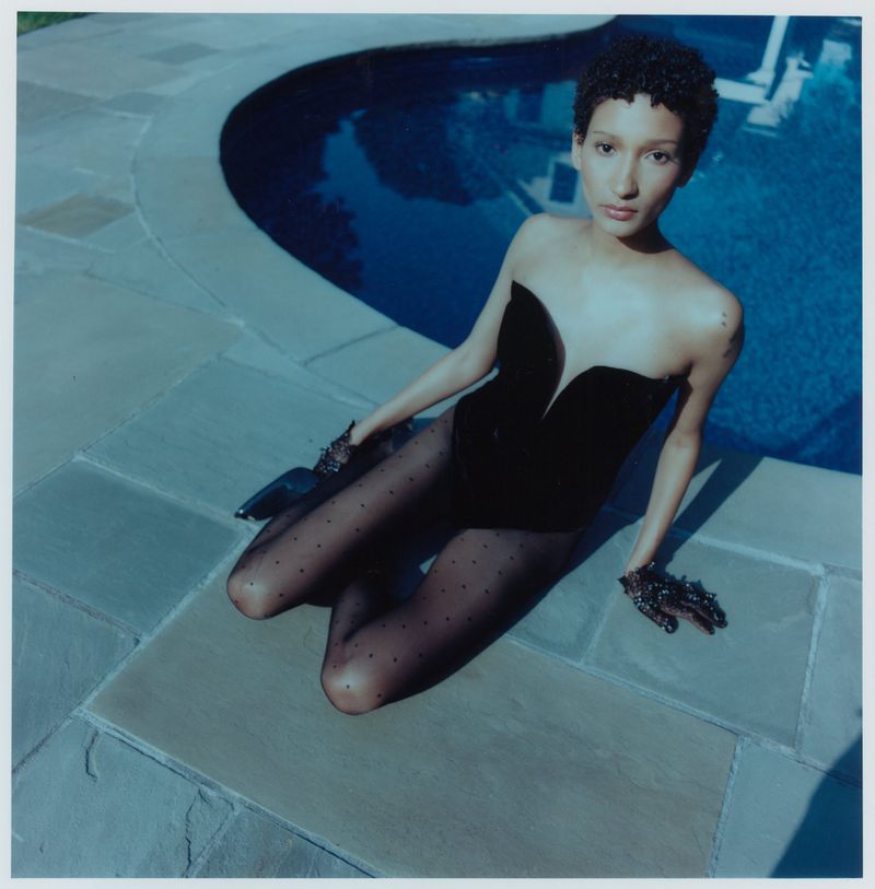 Young woman in strapless leotard and polka dot tights poses next to a pool on stone tiles, under natural lighting, in square format.