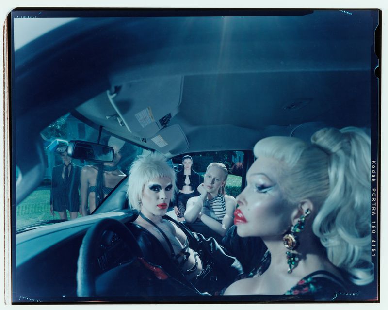 Indoor car scene with five people: two drag queens in front, three people in back; dark interior, cool lighting, and fashion elements.