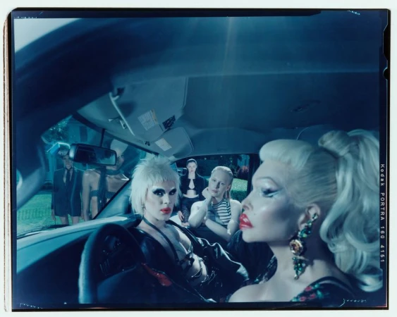 Indoor car scene with five people: two drag queens in front, three people in back; dark interior, cool lighting, and fashion elements.