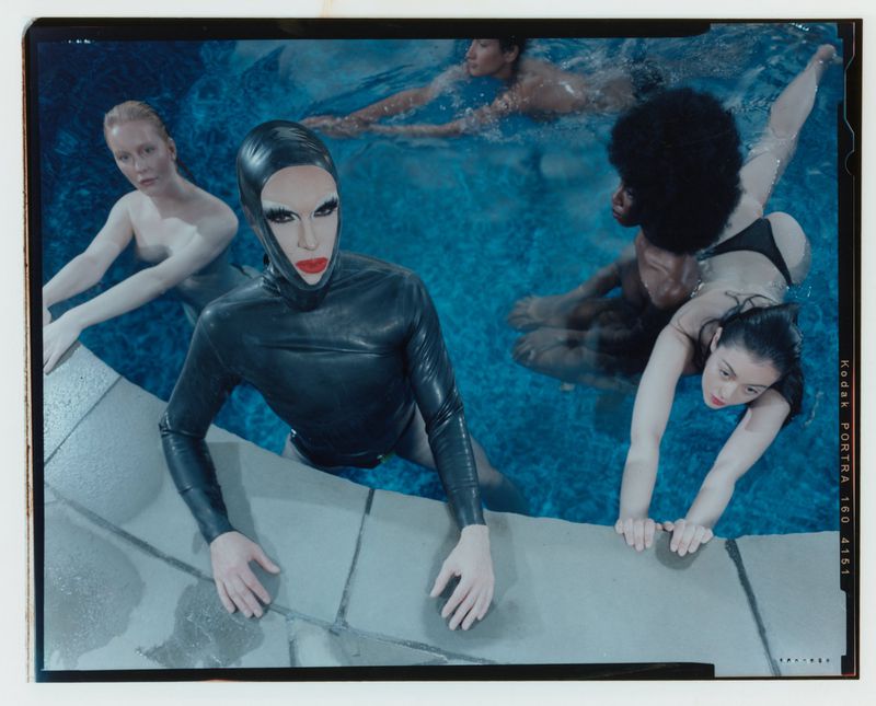 Group of five people in a swimming pool, one wearing a black latex suit with red lipstick, two are swimming and two are holding onto the pool edge.
