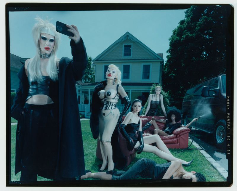 Group of people in avant-garde fashion posing outside a house with a van; one person takes a selfie in outdoor fashion editorial.