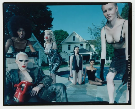 Group of six fashion models wearing swimsuits and dresses pose by a swimming pool in front of a suburban house on a sunny day.
