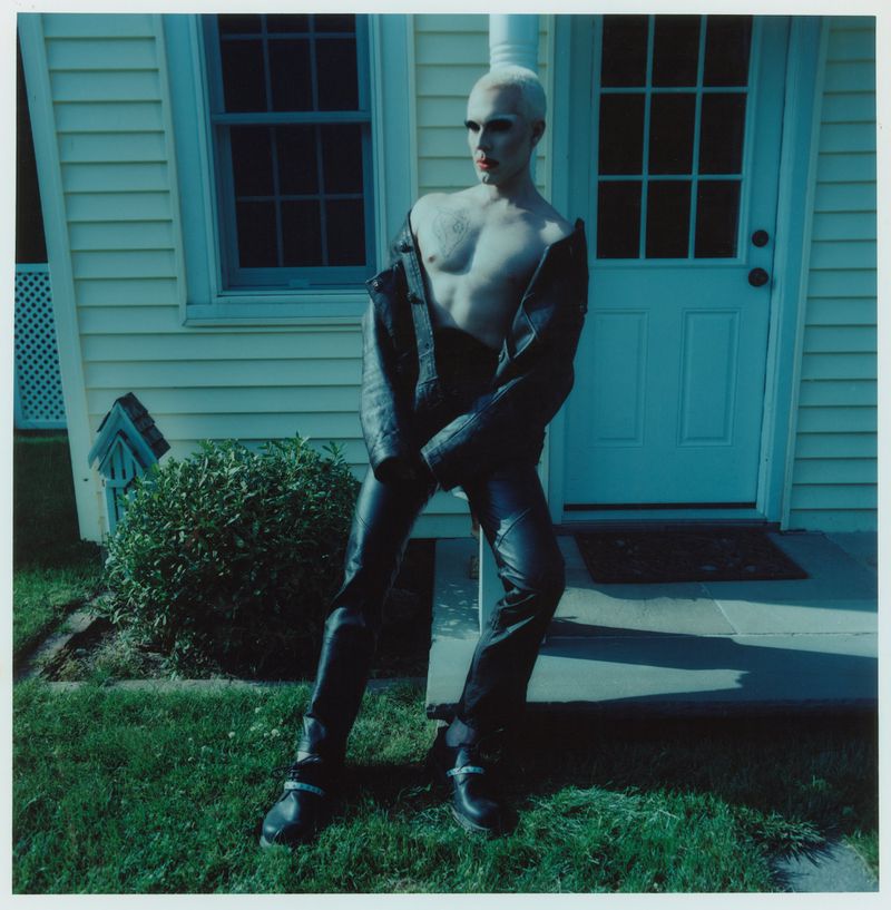 A shirtless person with light hair and red lipstick poses outdoors wearing leather pants and jacket, standing in front of a light yellow house.
