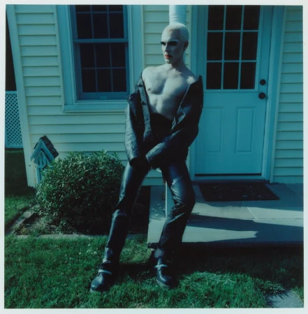 A shirtless person with light hair and red lipstick poses outdoors wearing leather pants and jacket, standing in front of a light yellow house.
