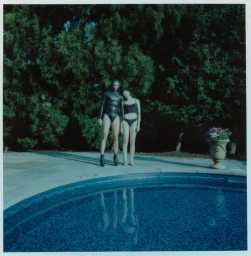 Two figures in black swim and latex attire standing beside an outdoor swimming pool against dense green pine trees.