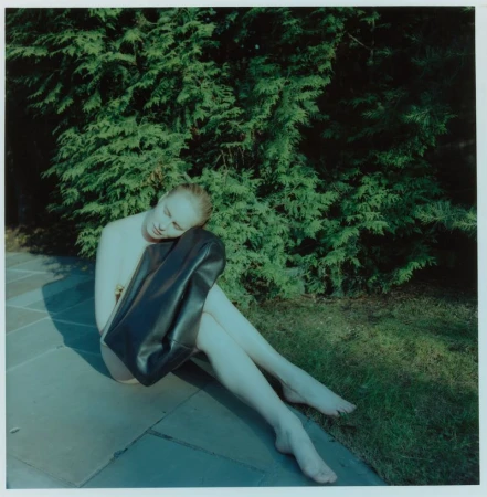 A seated model resting on stone paving against green shrubs, holding an oversized black leather tote bag.