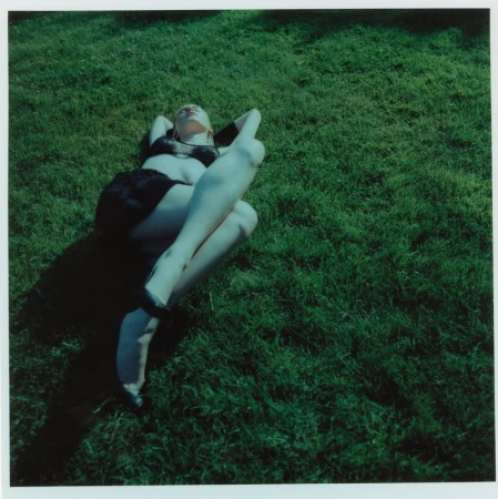 A woman lying on a green grass lawn wearing a black two-piece outfit and heels with arms behind her head under direct sunlight.