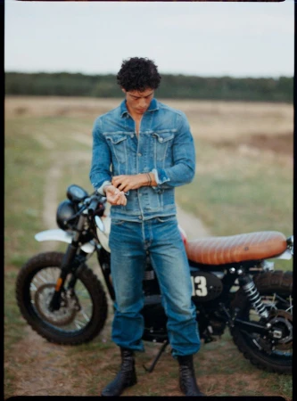 Man in denim jacket and jeans stands with motorcycle on a dirt road near a field and distant treeline under a bright sky.