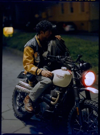 A couple on a motorcycle at night, embracing while on a Triumph Scrambler with a lit headlight, wearing a varsity jacket and boots.