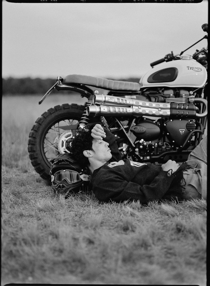Young man in sweater lies on grass with motorcycle and helmet in black and white, resting in outdoor, grassy landscape, profile view.