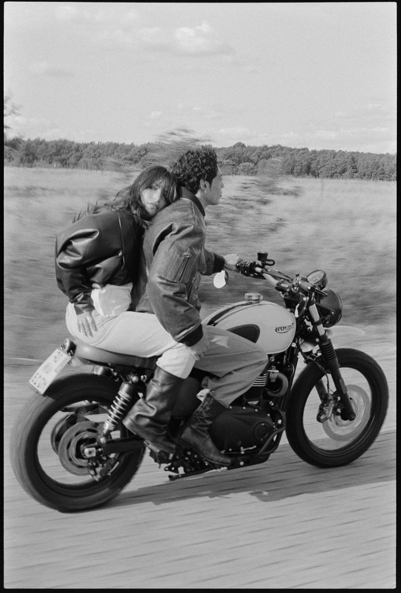 A couple riding a vintage Triumph motorcycle on a road with a blurred background of trees and sky, captured in black and white.