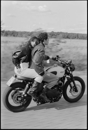 A couple riding a vintage Triumph motorcycle on a road with a blurred background of trees and sky, captured in black and white.