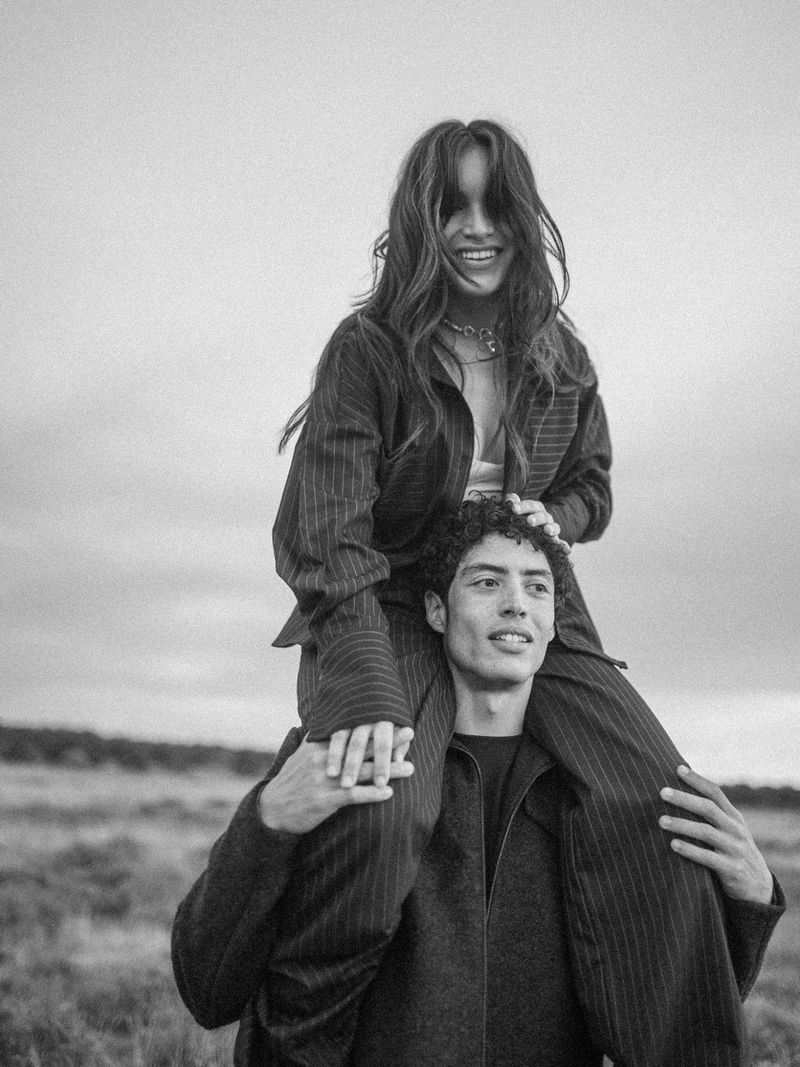 A woman sits on a man's shoulders in a monochrome outdoor portrait. The couple stands in a field under a cloudy sky, smiling.