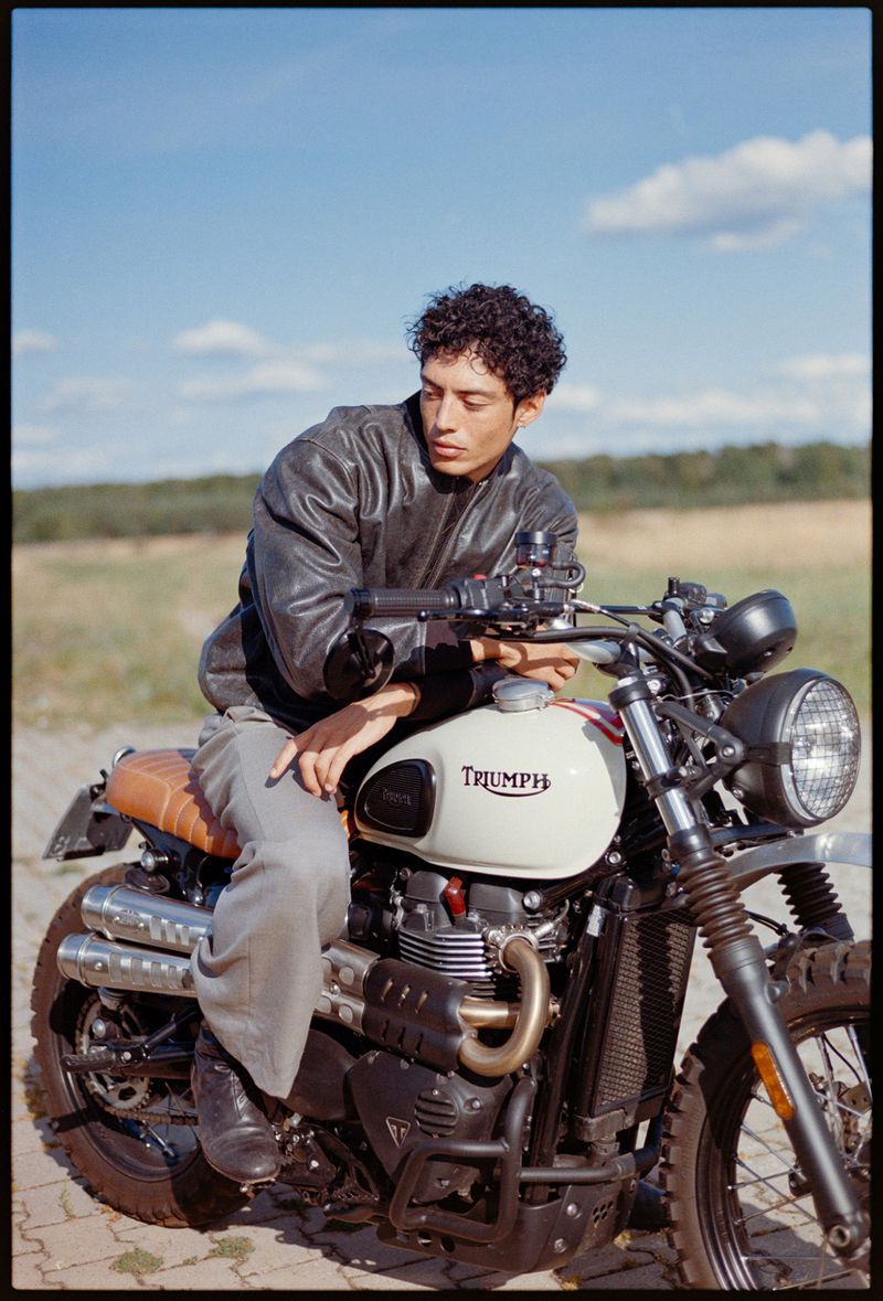 Man in leather jacket sitting on a Triumph motorcycle outdoors on a sunny day, parked on a brick surface, with blue sky in the background.
