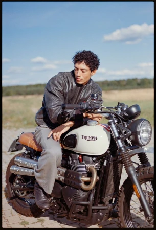 Man in leather jacket sitting on a Triumph motorcycle outdoors on a sunny day, parked on a brick surface, with blue sky in the background.