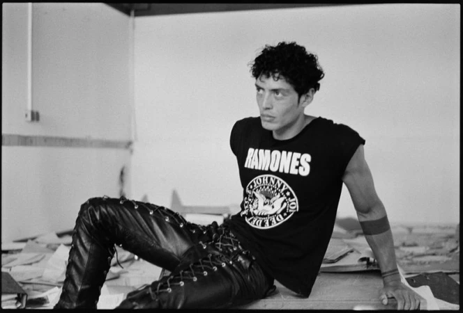Adult male with curly hair, seated and looking to the side. Wearing a Ramones t-shirt and laced leather pants in black and white.