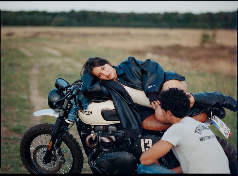 Woman in leather jacket rests on a Triumph motorcycle with a man seated nearby outdoors in a field, capturing a casual, intimate moment.