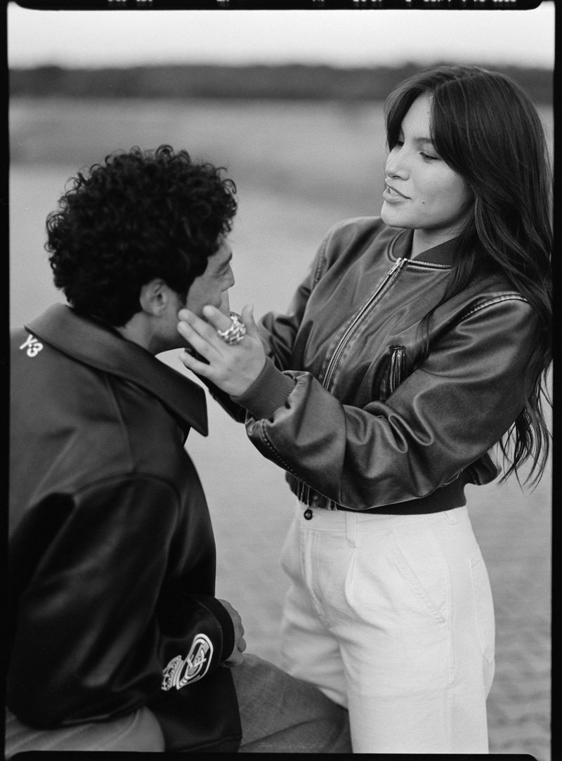 Black and white portrait of a woman in a leather bomber jacket touching a seated man's cheek outdoors.