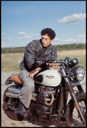 Model Cristiano Palmerini wearing a black leather jacket seated on a Triumph motorcycle outdoors under a blue sky.