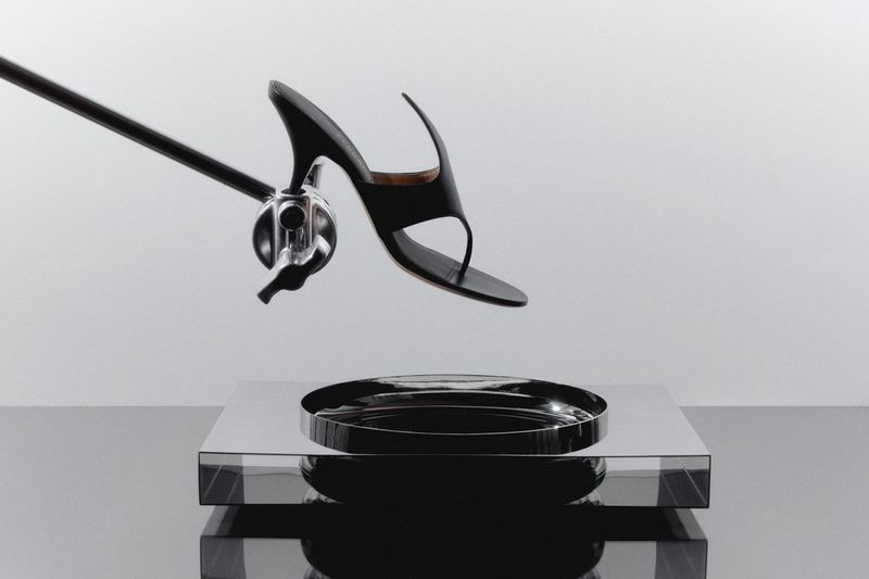 Elevated shot of black high-heeled sandal suspended above a reflective silver tray and surface, supported by a chrome arm and clamp.