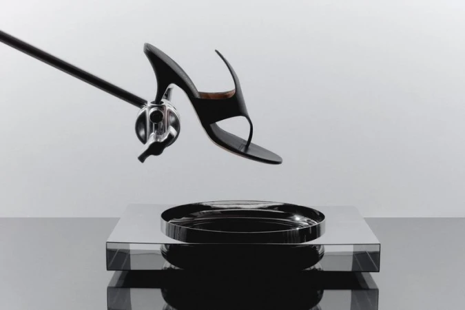 Elevated shot of black high-heeled sandal suspended above a reflective silver tray and surface, supported by a chrome arm and clamp.