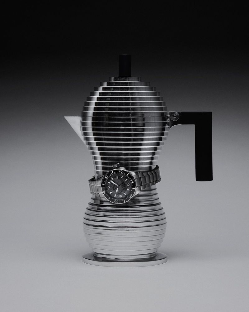 Studio shot of an aluminum moka pot with horizontal grooves. A silver wristwatch with a black face encircles the pot's waist.